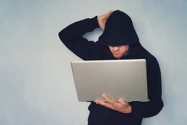 stock image thoughtful Hacker with his computer thinking on blue background.. hacker in a black hood with a gray modern ultrabook. identity theft.