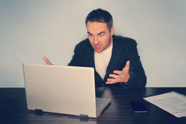 Frustrated young businessman looks at laptop screen screaming in anger at workplace in office. Unexpected high bill, unpaid debt, failing financial report tax delinquency, breach of contract concept.