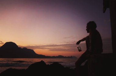A man drinks a strong whiskey and looks at the sunset. Lonely sad guy with a bottle of alcohol. The concept of loneliness and waiting. Young Man with a Beer sit at Seaside on Sunset Background clipart