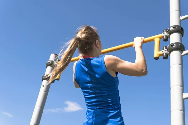 Girl on street workout. She pull-ups herself up on bar on sports ground in park. Photo from the back. Man is unrecognizable.
