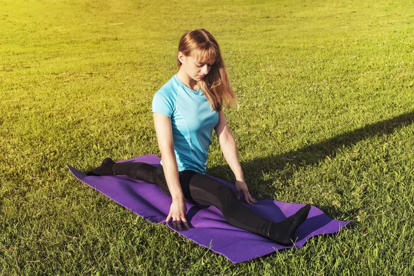 A dark-haired woman coach in a sporty short top and gym leggings makes a wide twine on the rug for yoga hands are raised upwards on the yoga mat, hands are raised upwards, on a summer day in a park