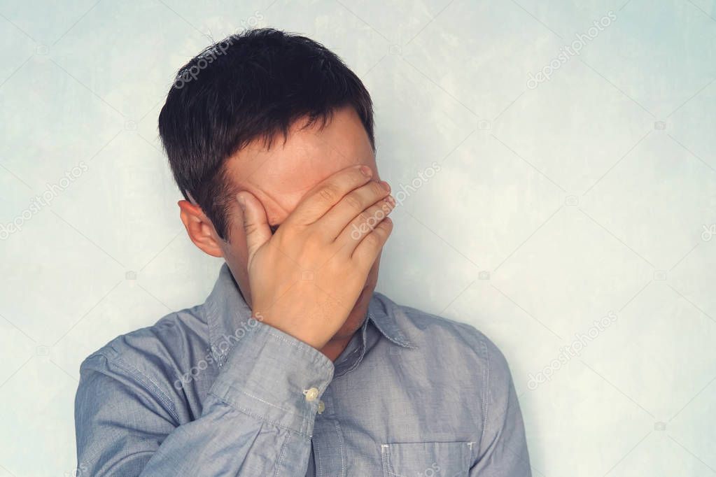 a young guy in a blue shirt is holding his head. the concept of fatigue. Close your eyes to the problem. Portrait of a handsome man covering his eyes with hands on a blue background