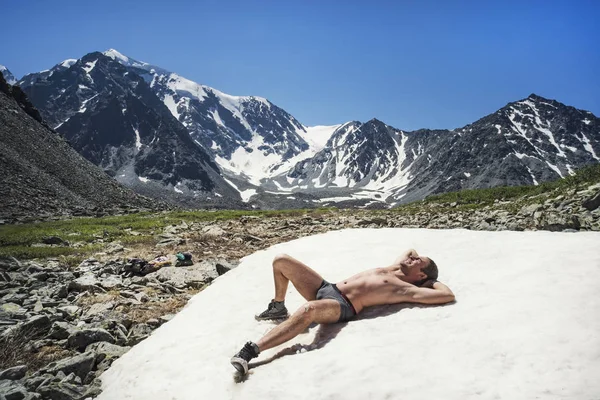 naked man on ice mountain against sky. Young guy lying on snow in mountains, sunbathing enjoying rest. Altai, Siberia, Russia. Russian entertainment in snow mountains in summer. harden, temper, harden