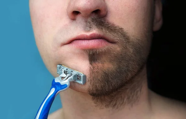 A man shaves his face without cream or foam, experiencing pain and suffering. Half face shaved half overgrown with a thick beard.