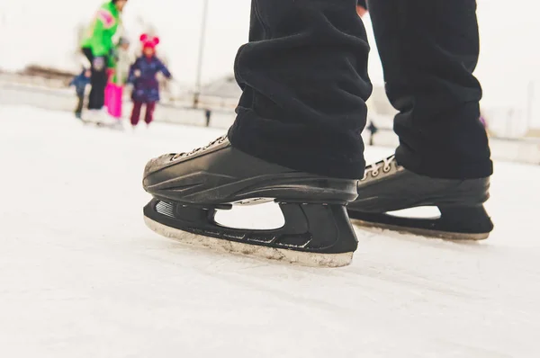 Men's feet in ice skating rink in black skates on the background of entertaining people who skate on the rink in the open air.