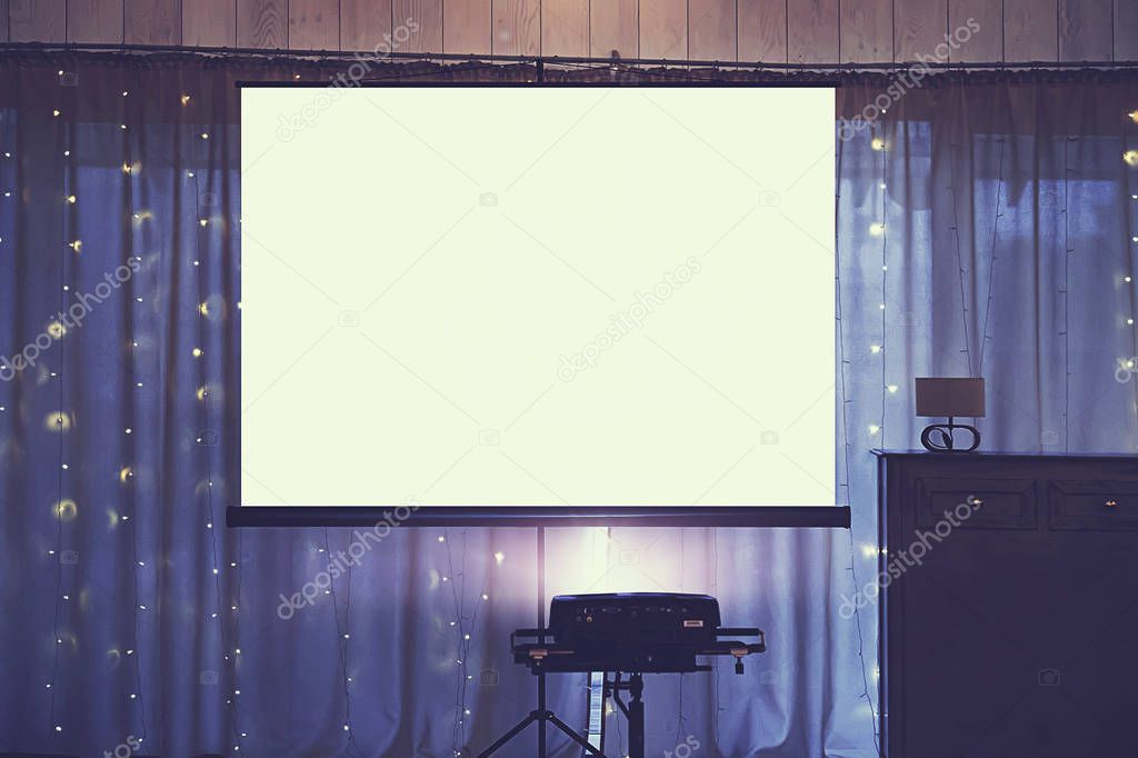Front view of decorated wedding room with empty white projector screen in the centre. Equipment for video and slideshow projection at a festive event. Banquette hall with a board for projection