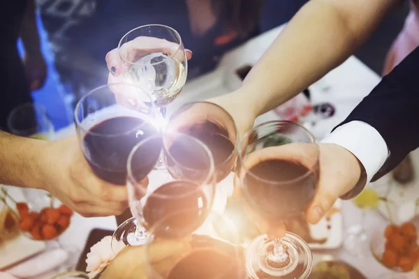 Lens flare short of male and female hands with filled glasses of wine above the restaurant table. Drinking toasts and clinking tumblers at a dinner party. Drinking wine at a banquet. Feast an event.