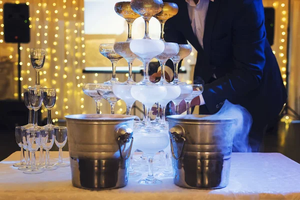 Man in suit fills the 4-tier champagne tower with sparkling wine at the illuminated banquet hall background. The glasses tower with champagne is on a table at festive occasion background. Cascade.