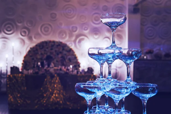 Awesome shot of glittering 3-tier champagne tower at the decorated banquet hall background. Beautifull built-in set of coupe glasses at a wedding room. Wedding champagne tower is ready for pouring