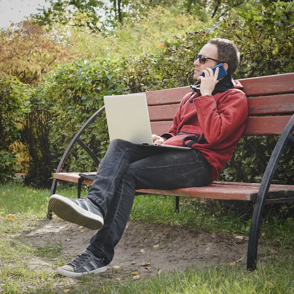 Homme travaillant à l'extérieur toujours connecté avec son smartphone et son ordinateur portable fidèle. jeune homme, pigiste travaillant en plein air à l'automne Park — Photo