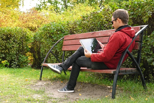 Un jeune homme en vêtements décontractés dans une veste chaude travaillant à l'extérieur dans le parc d'automne, lire un livre sur ultrabook. adolescent recherche contenu multimédia ensemble sur la ligne avec ordinateur portable assis sur le banc dans un parc — Photo