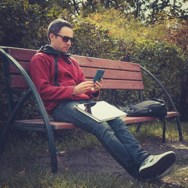 Jeune hipster gars vérifier notification sur téléphone mobile pendant le travail à distance sur ordinateur portable, assis dans le parc en été day.Handsome homme bavarder en ligne sur smartphone pendant le webinaire sur netbook — Photo