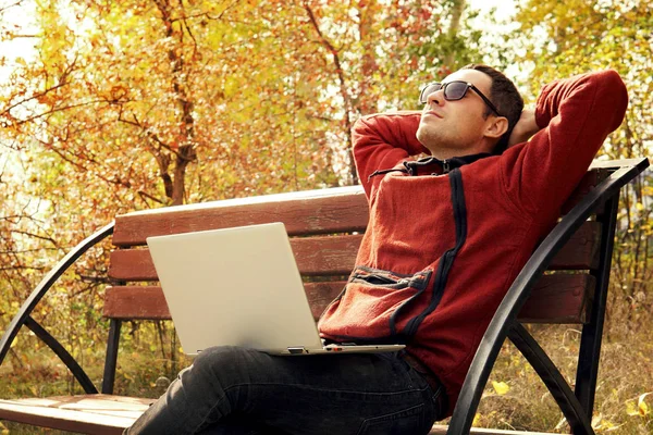 Счастливый взрослый мужчина с ноутбуком, сидя на заборе у озера. Молодой он специалист отдыхает от работы. Удаленная работа на открытом воздухе. Студента переписывают в Интернете — стоковое фото