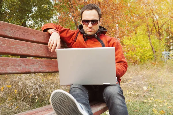 Jeune homme travaillant sur son ordinateur portable se connecter à Internet sans fil dans la zone rurale. La connexion Internet permet aux gens de travailler n'importe où. Programmeur freelance assis à l'ordinateur portable sur un banc dans le parc à l'extérieur . — Photo