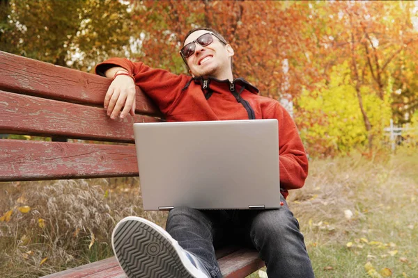 Молодой предприниматель празднует успех бизнеса, читая хорошие новости на ноутбуке в парке. Парень-фрилансер смеется, держа в руках ультрабук. Рабочий в повседневной одежде. Удаленная работа вне офиса — стоковое фото