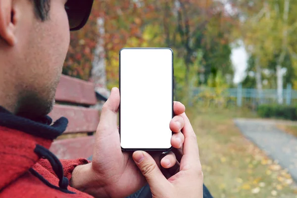 Joven mano sosteniendo teléfono inteligente con pantalla blanca al aire libre contra la acera de otoño. chico mira la pantalla de un teléfono inteligente vacío a la luz del día. usar el smartphone al aire libre . —  Fotos de Stock