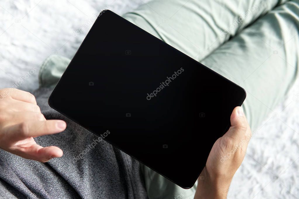 Close up of a young man comfortably lying on a bed with a tablet and touching the screen. Halfshot of a man sitting with black screen tablet in hands. Man's index finger raised over the touchscreen.