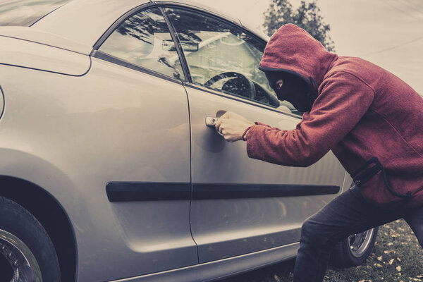 Side view of car being forced by a man in hoodie and mask. Thief tries to steal vehicle from a parking. Young male acts alone breaking the car door. Unknown person picklocks the automobile. Car fraud