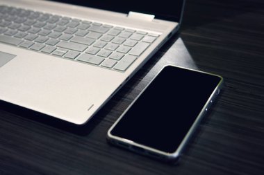 Kopyalama alanı ekranı ve ahşap masa arka plan üstünde laptop ile akıllı telefon. Boş siyah ekran bir mobil bilgisayarın yakınında tablo yaptı. İş yerinde dijital becerikli alet kullanımı. Telefon ekran mockup.