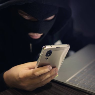 Siyah bir maske içinde erkek hacker smartphone ve dizüstü bilgisayar kullanır. Bir dolandırıcı siber suç tamamlar. Kişisel veri ve bankacılık ile sahte düzeni. Dolandırıcılık işlem yapmak için girişim. Cep telefonu hesabını dolandırıcılık