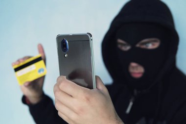 Hırsız scammer bir cep telefonu ve kredi kartı tutuyor. Banka kartı dolandırıcılığı ile a virüs içinde belgili tanımlık smartphone kavramı. bir haydut siyah yün ve siyah kukuleta.