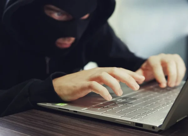 Fechar de um hacker mal intencionado em Balaclava preta a introduzir os dados num portátil. O hacker de computador comete cibercrime. Um homem sentado com tipos de rosto escondidos someting no laptop. Criminalidade na Internet — Fotografia de Stock