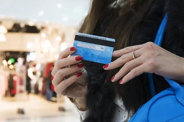 ぼやけているショッピング センターの背景で クレジット カードの裏面を押し赤のマニキュアで手入れの女性の手のクローズ アップ 若い女性は 銀行カードを使用して買い物をしています ブティックの女の子 — ストック写真