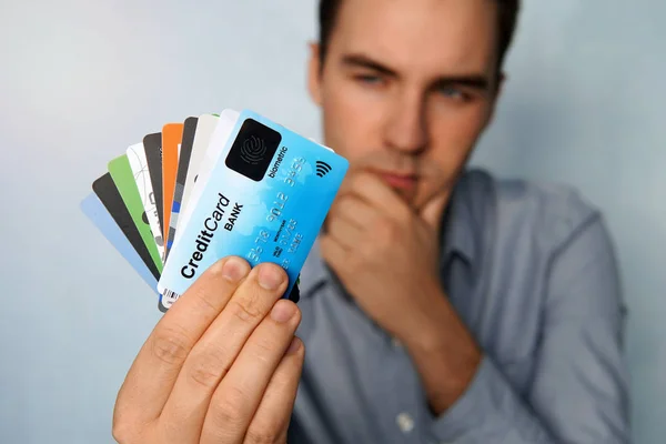 The young man is holding a stop of credit and debit cards in a pensive pose. The guy chooses a card to pay. businessman holds a large stack of credit cards in his hand and looks at them — Stock Photo, Image