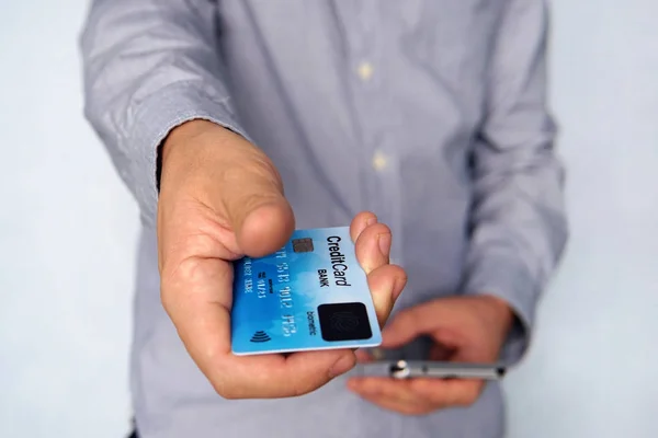 Cropped short of young man reaching out hand with blue credit card with fingerprint sensor and holding mobile in other hand. Businessman giving payment card with biometric scanner at blue background. — Stock Photo, Image