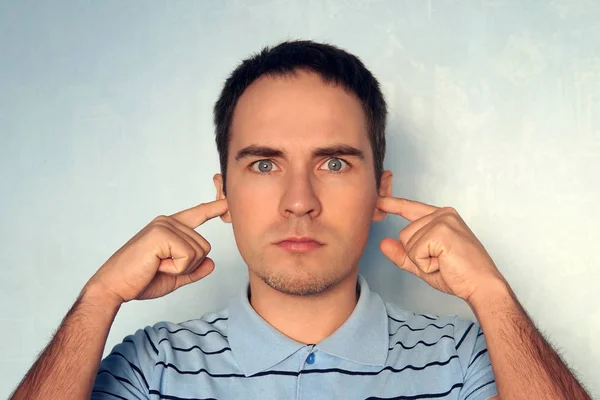 Handsome young male model plugs ears with displeased expression as hears annoying loud noise or roaring not far from him, over blue concrete wall. Not pleasant irritated sound.