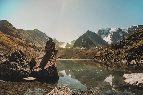 El joven viajero se sienta en una piedra y admira el hermoso lago de montaña en las montañas de Altai. Siberia Rusia. Un hombre con uniforme militar de camuflaje. Turista sentado en el acantilado sobre el lago . — Foto de Stock