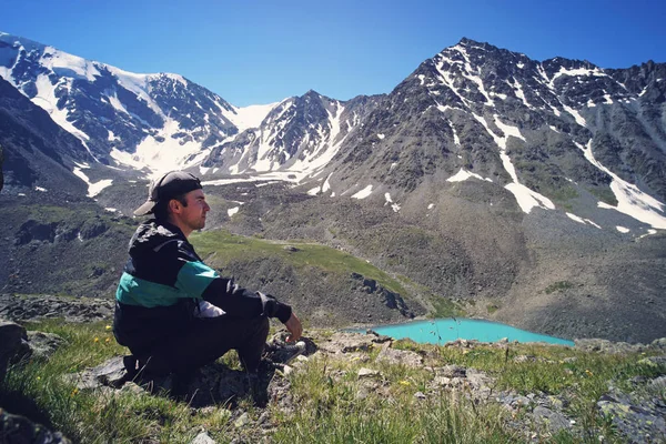 El joven en ropa deportiva negra está sentado en el borde del acantilado y mirando hacia el valle brumoso a continuación. Hermoso paisaje de fondo con altas montañas con picos nevados — Foto de Stock