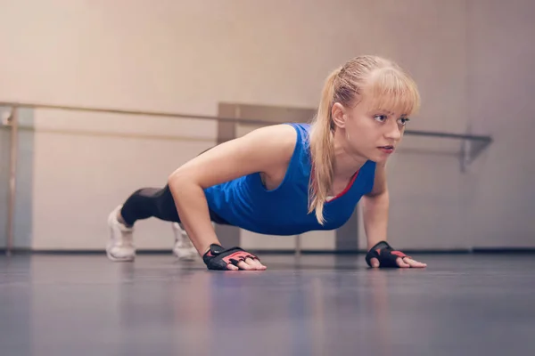Linda hermosa rubia con ojos azules en una camiseta deportiva azul haciendo ejercicios en el suelo. Chica atleta haciendo push-UPS en el gimnasio — Foto de Stock