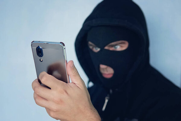 Young adult in black clothes with hidden face looks at smartphone screen. Ill-intended fraudster uses mobile. Fraudster calls. Mobile racket. Hacker hijacks by phone. Cellphone account fraud. Scam.