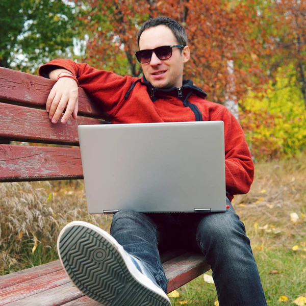 Joyeux gars communique par Internet dans la rue. Programmeur profiter avec ordinateur portable assis dans le parc d'automne. Un bel homme qui travaille sur son ordinateur portable. Travail gratuit à l'extérieur, indépendant à l'extérieur — Photo