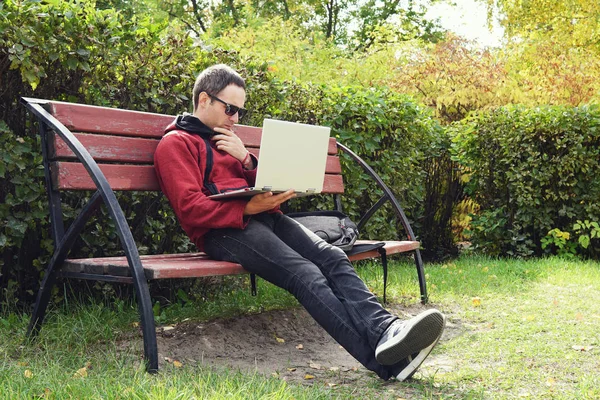 Jeune élégant message texte étudiant masculin sur ordinateur portable dans le parc à l'automne. Il travaille sur un ordinateur portable en plein air. Jeune homme réfléchi regardant l'écran de l'ordinateur portable.. Travail à distance — Photo