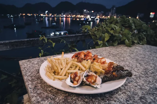 Plato con mariscos, mejillones, camarones, pescado, pulpo y papas fritas en la mesa contra el mar por la noche al aire libre. Vietnam ha Long Bay  . — Foto de Stock