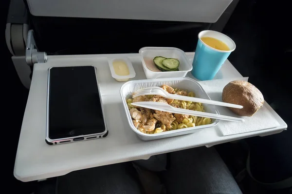 Varios alimentos de clase económica y un teléfono inteligente de pasajeros con una pantalla en blanco en la mesa plegable del avión. Comidas a bordo del avión. Cuchillería de plástico cuchillo y tenedor en un plato con comida . — Foto de Stock