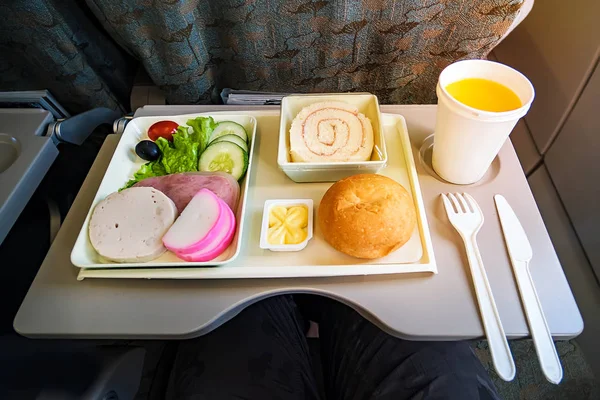 Bandeja de servicio de comidas en vuelo para clase económica, carne, fruta, ensalada, pepino, un vaso de jugo y mantequilla. cuchillo tenedor de plástico para mayor seguridad . — Foto de Stock