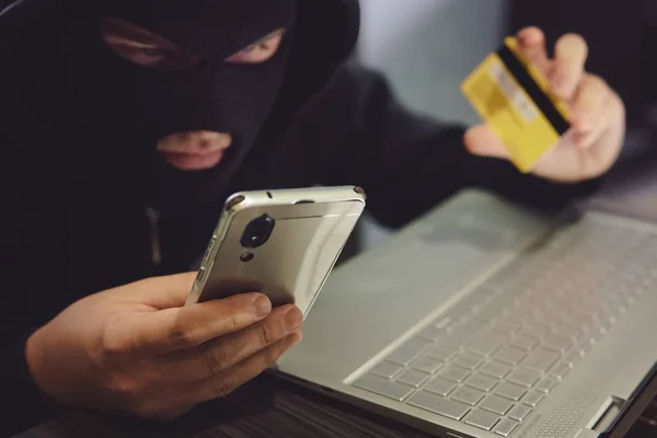 Male hacker in a robber mask uses phone, credit card and laptop in some fraudulent scheme. Cyber thief stole the personal data and credit card information. Hacker uses malware to steal user's money — Stock Photo, Image