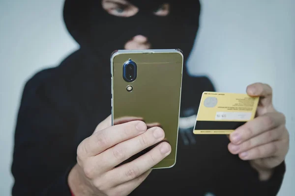 Cyber criminal in balaclava enters the information of a personal bank account. Credit card fraudulent scheme. Stealing cyber money using mobile. New ways of fraudulent transactions via online banking — Stock Photo, Image