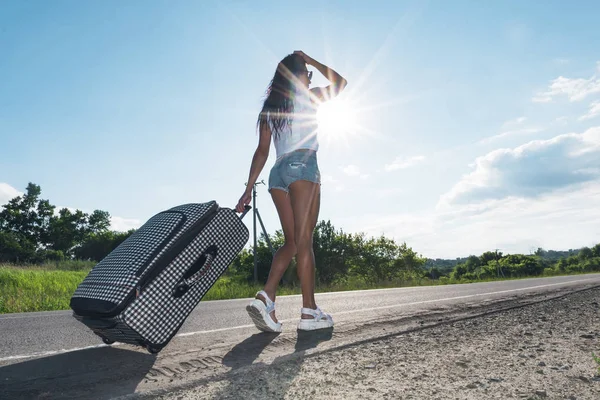 Девушка под ярким солнцем на дороге с багажом на заднем плане автомобиля. женщина едет по дороге с чемоданом. концепция приключений и путешествий в одиночку. Дорожные поездки. автостопом. Летние выходные . — стоковое фото
