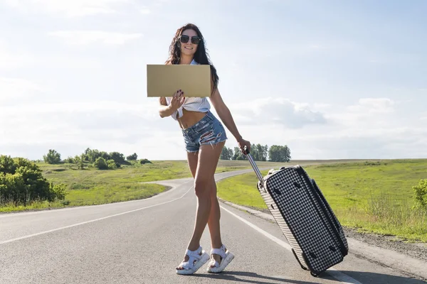 Девушка с туристическим чемоданом. автостопом по дороге и проведение картона, открытый, готов к лету, отдых и путешествия концепции. красивая, молодая сексуальная брюнетка — стоковое фото