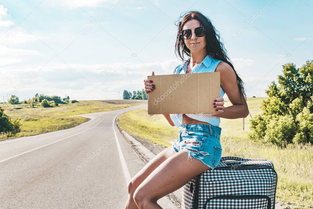 Pretty young woman hitchhiking along a road and waiting on a country road with her suitcase and empty cardboard plate. Beautiful girl in sunglasses hitchhiker. empty Board