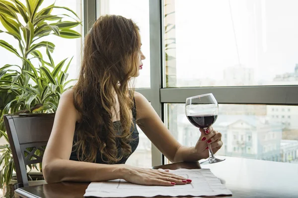 Una joven hermosa con un vestido negro se sienta en un restaurante y bebe vino de una copa. primer plano de la joven mujer que su mano sosteniendo con vino tinto de cristal como un mirando hacia fuera en la ventana. — Foto de Stock