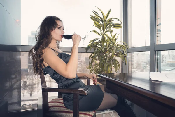 Νεαρή όμορφη γυναίκα κάθεται σε ένα τραπέζι σε ένα καφέ με αργά πίνοντας ένα ποτήρι κρασί κοιτάζοντας έξω από το πανοραμικό παράθυρο με μια όμορφη θέα της πόλης. — Φωτογραφία Αρχείου