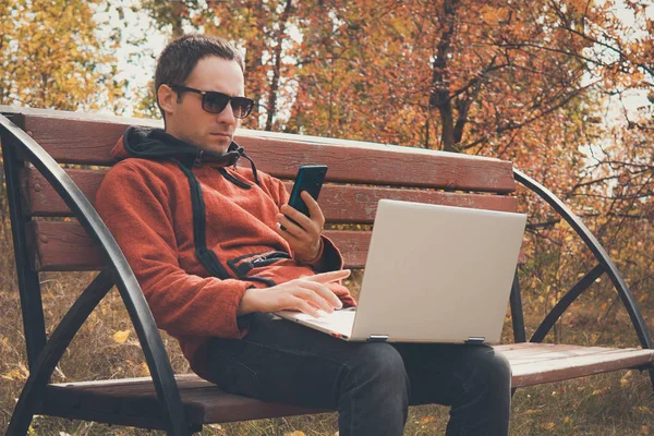 Jeune homme indépendant utilisant un téléphone mobile et un ordinateur portable pc.Adolescent parlant sur smartphone dans un parc urbain. gars avec ordinateur sans fil extérieur. Concept technologique. L'été indien. automne moelleux . — Photo