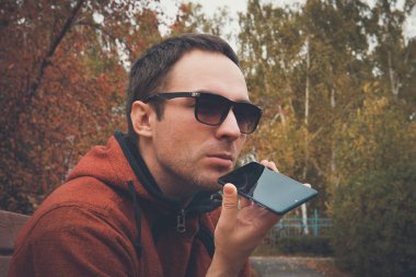 Genç yakışıklı adam yaz aylarında parkta duran, kaydedilen sesli mesaj veya ses dinlemek için bir cep telefonu kullanarak. adam mikrofona bir sesli mesaj kaydetmek için konuşuyor. Diktafonda kayıt