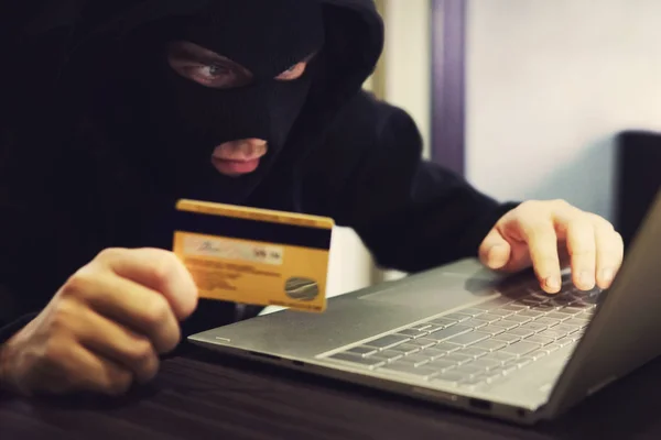 Man in robber mask and hood misappropriates personal bank data. Cyber fraudster attacks online banking system. Hacker makes financial fraud via internet. Digital crime with the use of payment card.