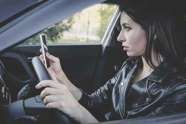 Vista de perfil de una mujer joven y atractiva mirando su teléfono inteligente mientras conduce un coche en un día cálido. Conductora en chaqueta de cuero revisando el celular al volante. Chica sostiene la rueda y lee móvil . — Foto de Stock